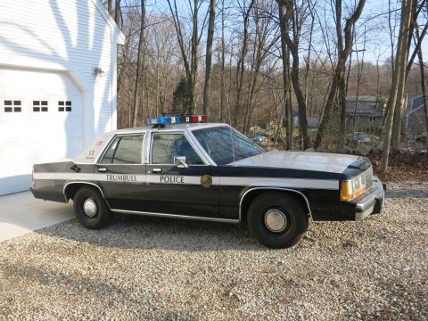 1991 Ford LTD Crown Victoria - black-and-white Trumbull Police Ford sedan with rooftop lightbar sits on a gravel driveway beside a white garage.