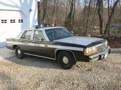 1991 Ford LTD Crown Victoria - Front three-quarter view of a black-and-white Trumbull Police Ford sedan, unit 13, parked on a gravel drive by bare woods.