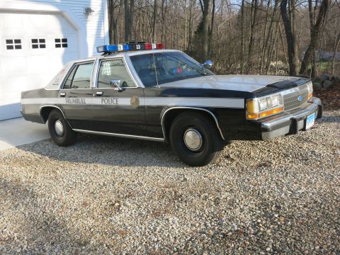 1991 Ford LTD Crown Victoria - black-and-white Trumbull Police Ford sedan with grille push bar and lightbar, unit 13, on a gravel driveway.