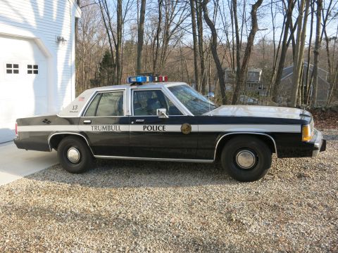 1991 Ford LTD Crown Victoria - Driver-side profile of a black-and-white Trumbull Police Ford sedan, unit 13, parked on gravel near a garage.