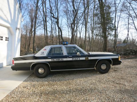 1991 Ford LTD Crown Victoria - Rear three-quarter view of a black-and-white Trumbull Police Ford sedan, unit 13, on a gravel drive beside a white garage.