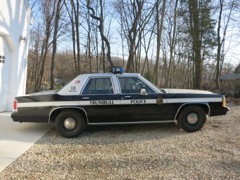 1991 Ford LTD Crown Victoria - Side view of a black-and-white Trumbull Police Ford sedan, unit 13, with U.S. flag decal, parked on a gravel driveway.