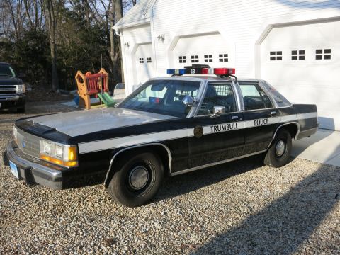 1991 Ford LTD Crown Victoria - Front three-quarter view of a black-and-white Trumbull Police Ford sedan, unit 13, parked on gravel before two white garage doors.