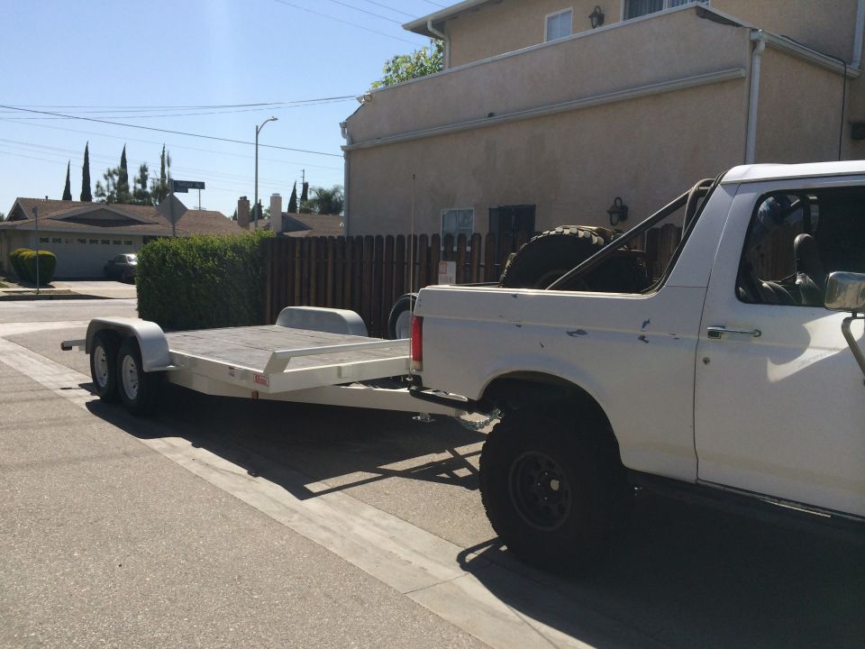White pickup truck with a roll bar towing an empty flatbed trailer on a residential street