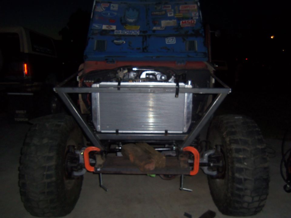 Off-road vehicle front view with large radiator, custom roll cage, and oversized tires. Another vehicle is partially visible to the left