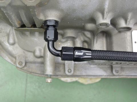 Close-up of an engine block with a black braided hose and fittings attached. Text on hose fitting reads 'Jiffy