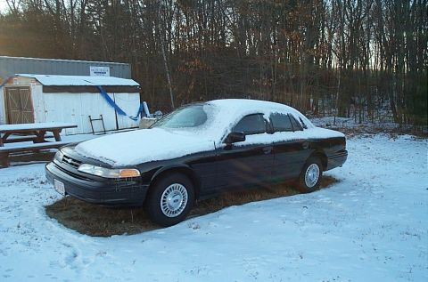 1991 Ford Crown Victoria LTD - A black sedan sits covered in snow, parked in a snowy yard with trees and buildings behind it.