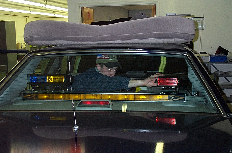 Dark colored car interior viewed from rear, showing a person installing red and blue emergency lights on the rear window shelf