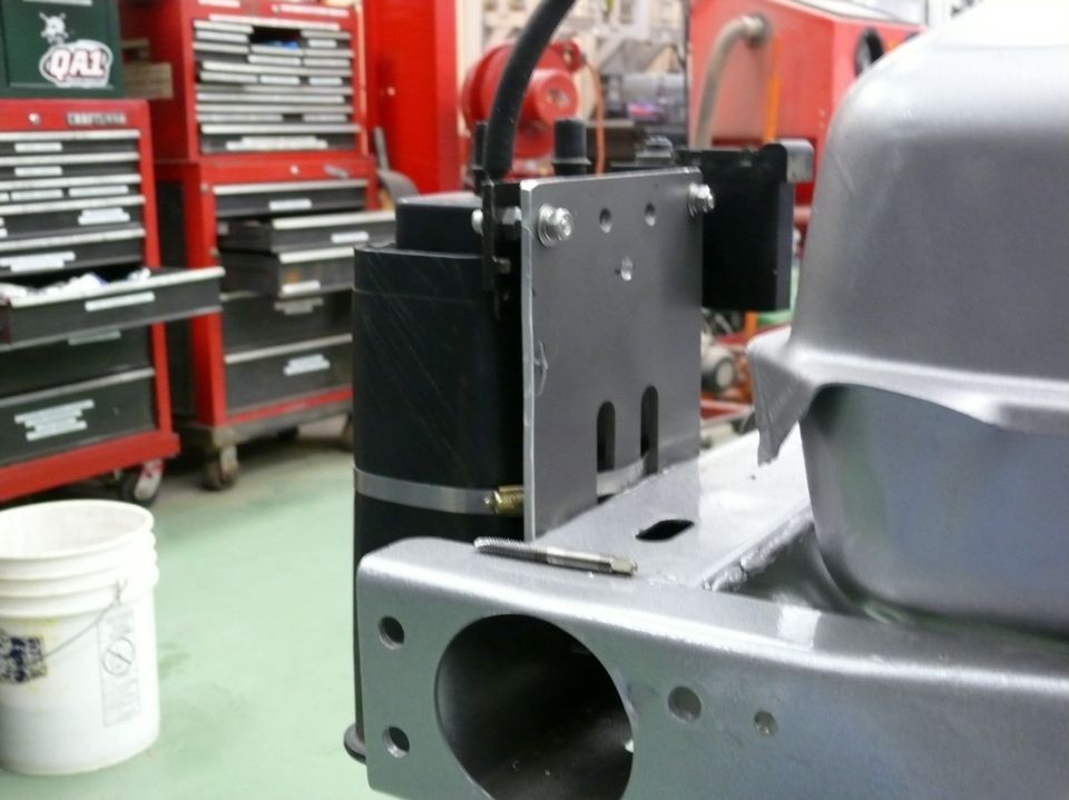 Close-up of a black cylindrical part mounted to a metal bracket, with a silver fuel tank visible on the right. Tools and red tool chests are in the background