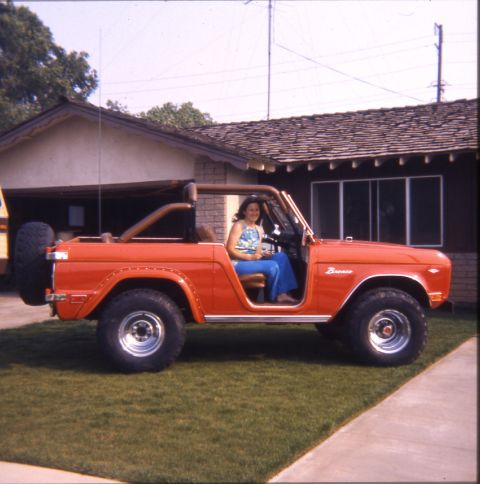 1996 Ford Bronco - A woman sits in a classic orange Ford Bronco parked in front of a house.