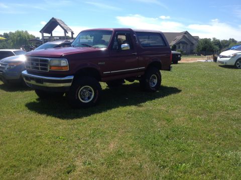 1996 Ford Bronco - A maroon 1996 Ford Bronco is parked on a grassy field under a blue sky.