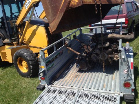 1996 Ford Bronco - A backhoe is loading a trailer with engine and axle parts, while a Ford Bronco is parked nearby.
