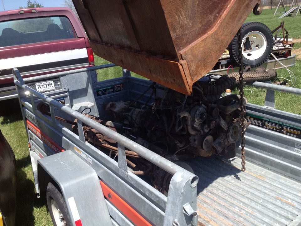 Engine being lowered into a trailer by a rusty bucket, with a maroon truck in the background