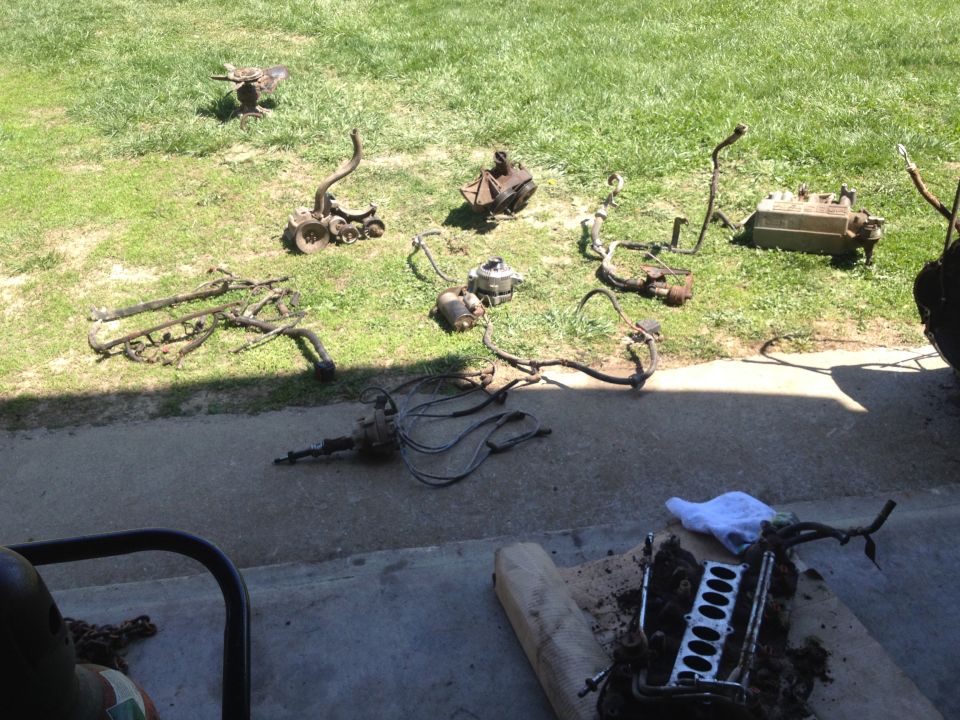 Disassembled engine components laid out on grass and concrete, including a transmission, exhaust manifold, and cylinder head