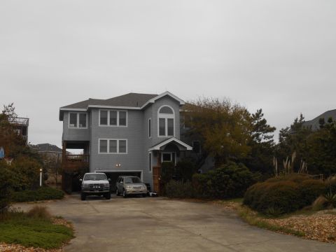 1994 Ford Bronco - Two vehicles are parked in the driveway of a large, gray, multi-story house on an overcast day.