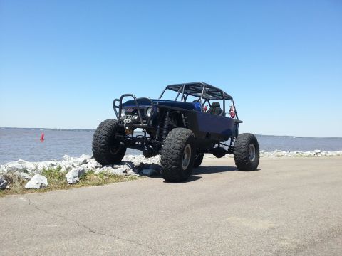 1967 Ford Mustang - A custom-built buggy with a roll cage and oversized tires sits on a paved path near a rocky shoreline under a clear blue sky.