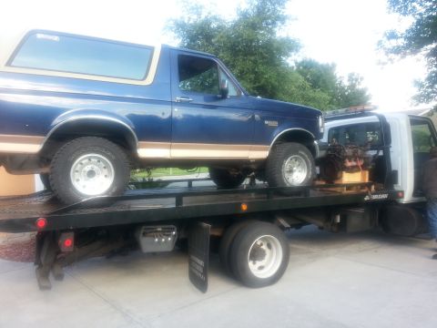1994 Ford Bronco - A blue Ford Bronco is loaded onto a flatbed tow truck, with an engine visible on a pallet behind it.