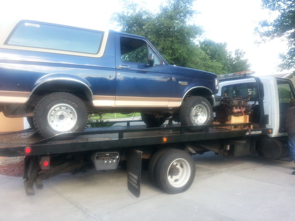 Blue Ford Bronco with tan trim on a flatbed tow truck, side view. Engine on a pallet behind it