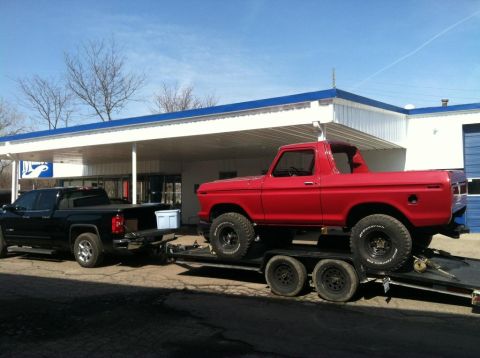 1978 Ford Bronco - A red 1978 Ford Bronco is loaded onto a trailer, towed by a black pickup truck in front of a building.