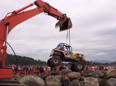 1982 Ford Bronco - An excavator lifts a heavily modified off-road jeep during an event with spectators in the background.