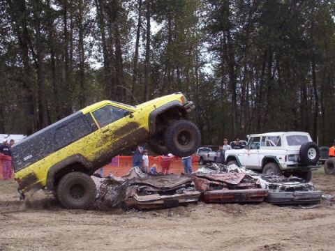 1982 Ford Bronco - An off-road event features a yellow Jeep Cherokee on top of crushed cars, while a white Ford Bronco waits nearby.
