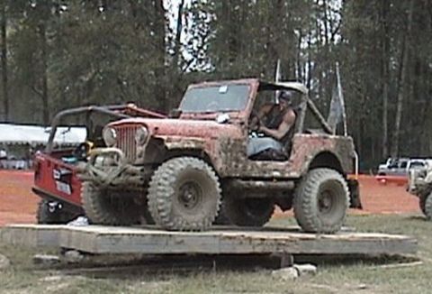 1982 Ford Bronco - A driver sits in a red Jeep on a wooden platform during an off-road event.