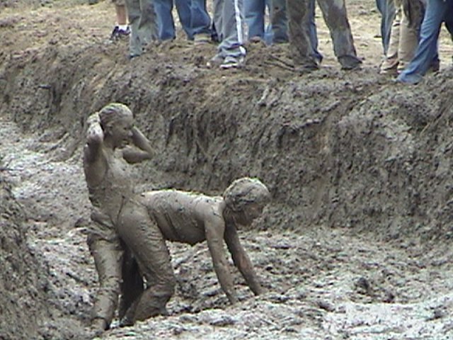 Two people covered in mud in a muddy trench, with spectators standing above