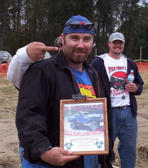 1982 Ford Bronco - Man holds a '2003 Roll Over Award' a lifted truck, with another man in the background.
