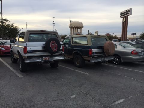 1996 Ford Bronco - Two Ford Broncos are parked side-by-side in a parking lot under an overcast sky.