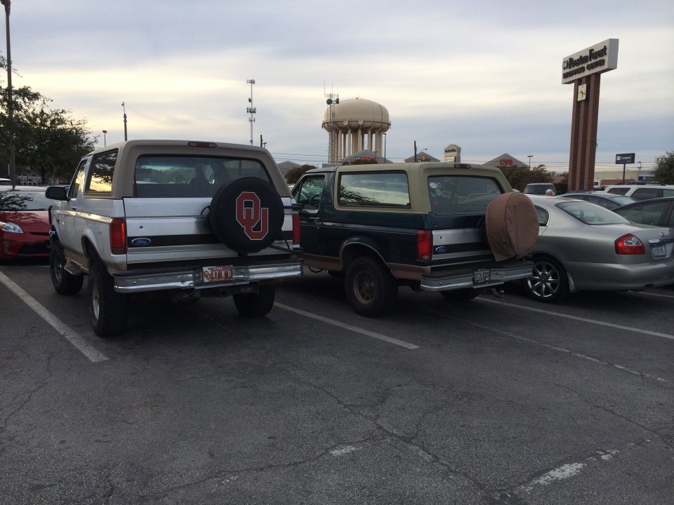 Two Ford Broncos parked in a lot, one silver and one dark blue, both with spare tires mounted on the rear
