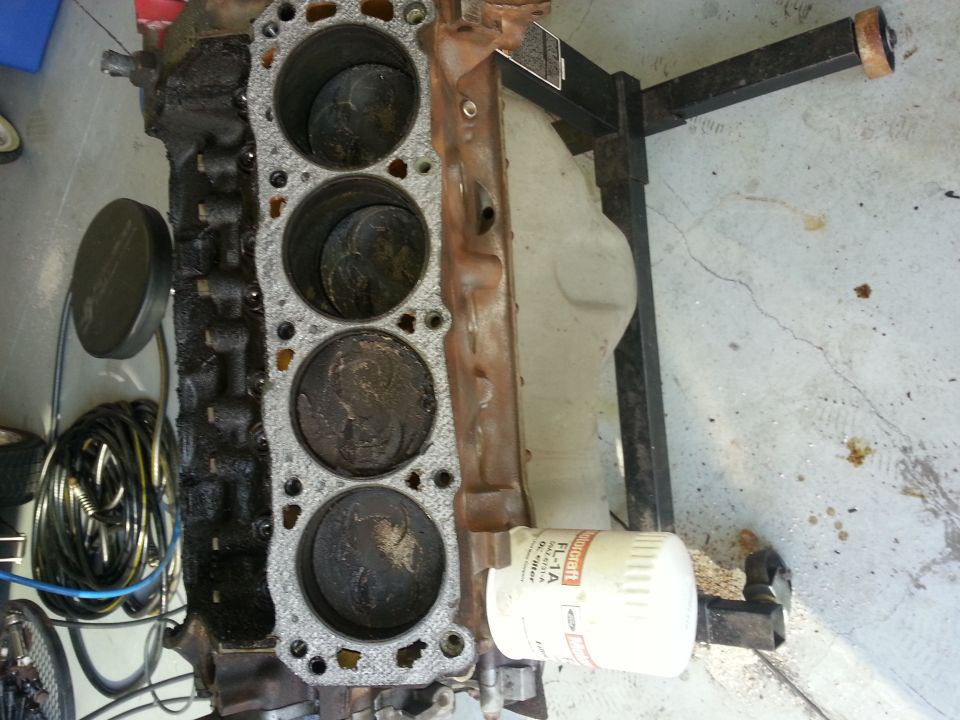 Engine block with four cylinders visible, pistons inside, and an oil filter attached. Mounted on an engine stand