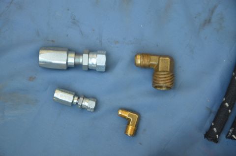 1979 Ford Bronco - Various hydraulic fittings and brass elbows are laid out on a blue surface, with a portion of a braided hose visible on the right.