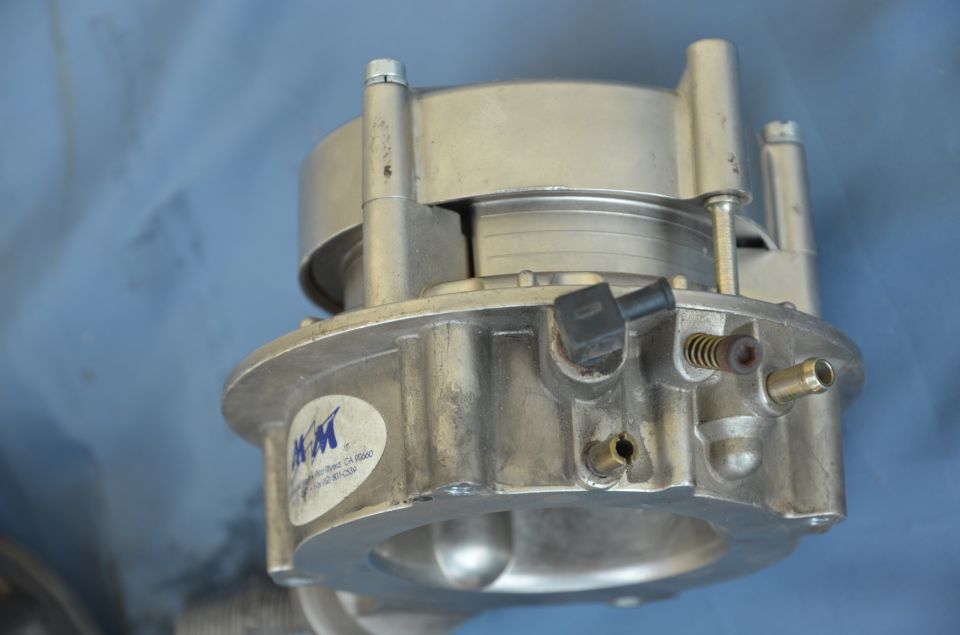 Close-up of a metal throttle body with a blue and white KM sticker. Part of a SuperMotors.net watermark is visible