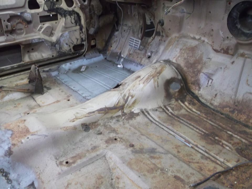 Car interior floor with rust and exposed metal panels, showing door frame and pedals