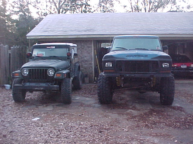 Two lifted dark 4x4s, a Jeep Wrangler and a full-size SUV with tube bumper, parked in front of a garage