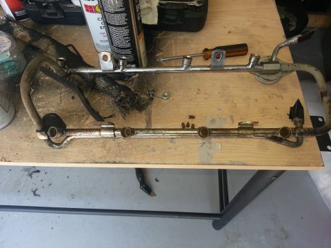 1994 Ford Bronco - A disassembled fuel rail sits on a workbench, surrounded by various automotive tools and cleaning supplies.