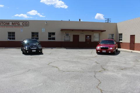 2003 Ford Crown Victoria - black sedan and a dark red Mercury sedan sit parked in a cracked lot before a tan 'Custom Mfg. Co.' building.