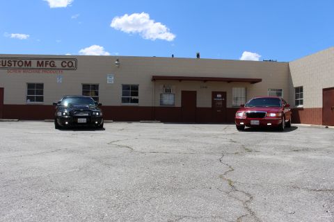 2003 Ford Crown Victoria - black sedan and a red Mercury sedan park outside a 'Custom Mfg. Co. Screw Machine Products' building under a blue sky.