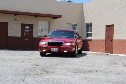 2003 Ford Crown Victoria - red Mercury sedan with EVIL MGM plate sits at an angle in a cracked parking lot beside a tan building.