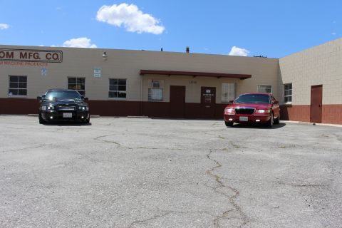2003 Ford Crown Victoria - black sedan and a red Mercury sedan park well apart in a wide cracked lot in front of a tan industrial building.