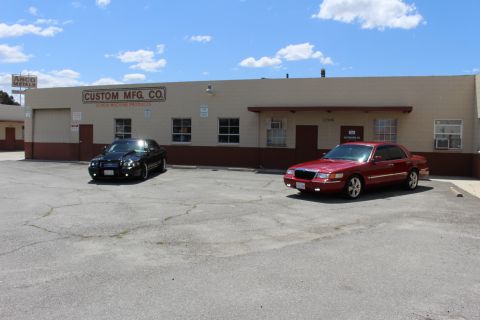 2003 Ford Crown Victoria - black sedan and a red Mercury sedan sit parked before the 'Custom Mfg. Co.' building with an Asco Mills sign at left.