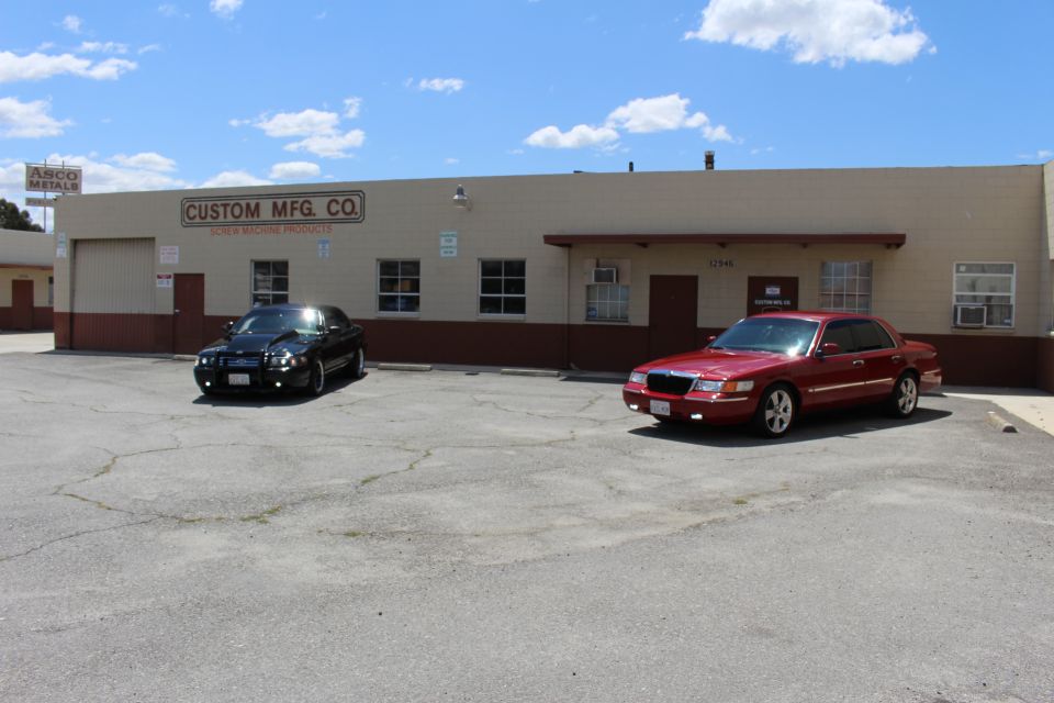 Black sedan and red Mercury sedan parked before Custom Mfg Co and Asco buildings