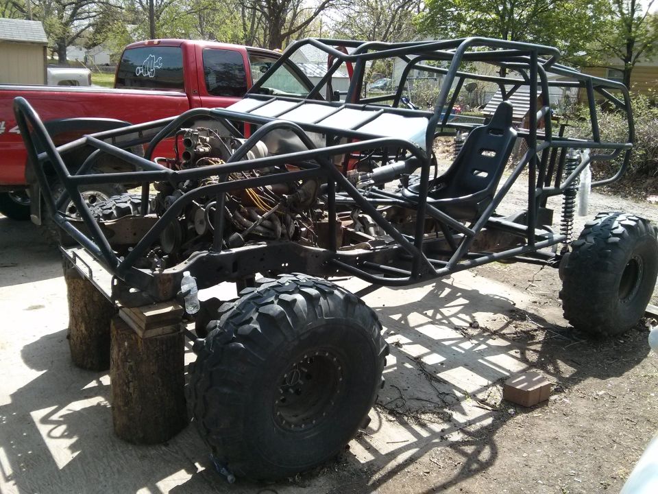 Custom buggy frame with large tires and exposed engine components, parked next to a red pickup truck