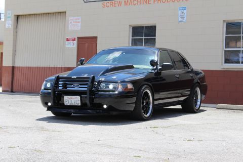 2003 Ford Crown Victoria - lowered black Ford sedan with a chrome brush guard, black wheels and 'EVIL VIC' plate parked outside an industrial building.