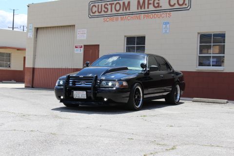 2003 Ford Crown Victoria - Front three-quarter view of a lowered black Ford sedan with brush guard and 'EVIL VIC' plate before the Custom Mfg. Co. building.