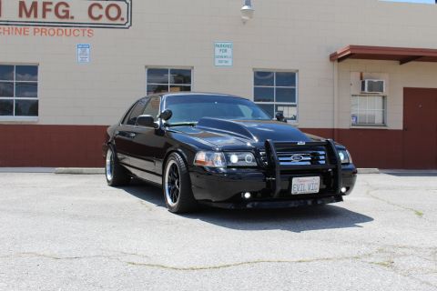 2003 Ford Crown Victoria - blacked-out Ford Crown Victoria with a chrome-trimmed push bumper, dark wheels and an 'EVIL VIC' license plate sits in a paved lot beside a…