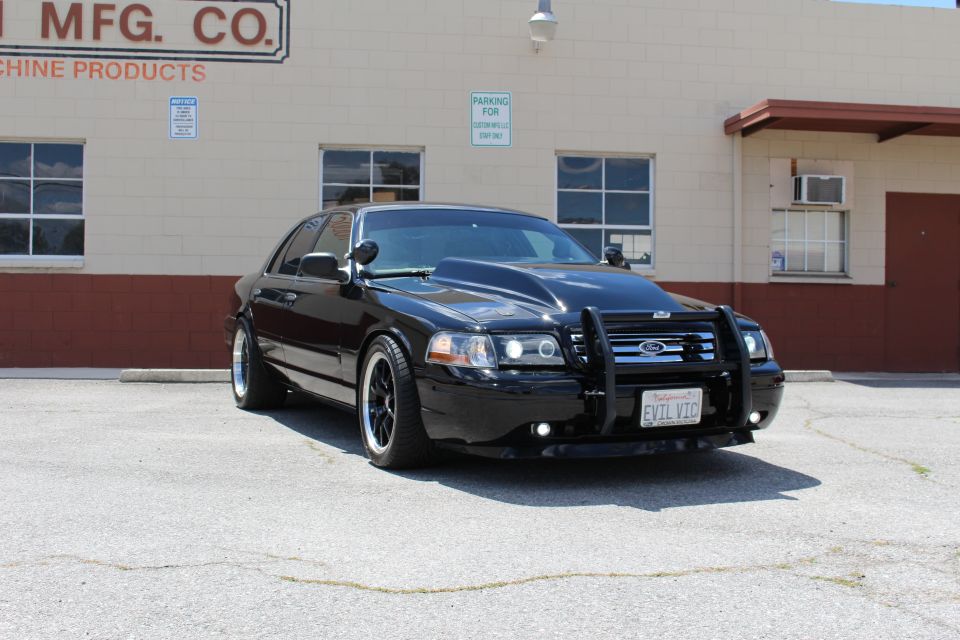 Black Ford Crown Victoria sedan with push bar and EVIL VIC plate parked by an MFG Co. building