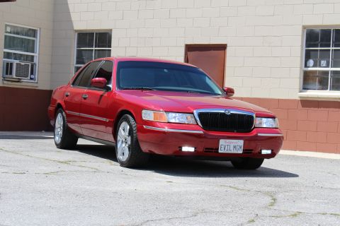 2003 Ford Crown Victoria - glossy red Mercury Grand Marquis with chrome wheels and an 'EVIL MGM' plate sits angled in a lot beside a tan and maroon building.