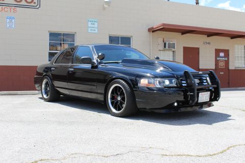 2003 Ford Crown Victoria - blacked-out lowered Ford Crown Victoria with a chrome push bumper and dark wheels sits angled in a paved lot beside a tan industrial buildi…