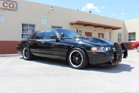 2003 Ford Crown Victoria - Side-front view of a lowered black Ford Crown Victoria with a folded push bar over the hood and polished-lip black wheels, parked at an ind…